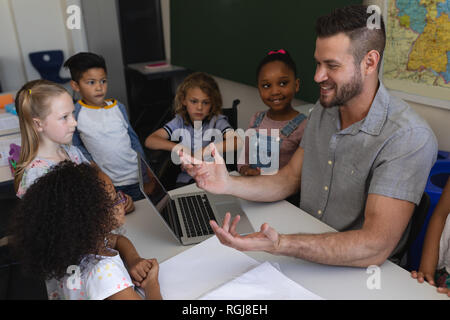 Front view happy Caucasian male school teacher teaching schoolkid at desk all kids look at him in classroom of elementary school Stock Photo