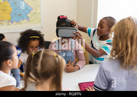 Front view schoolkids using virtual reality headset one of them help his classmate to put it on her head in classroom of elementary school Stock Photo