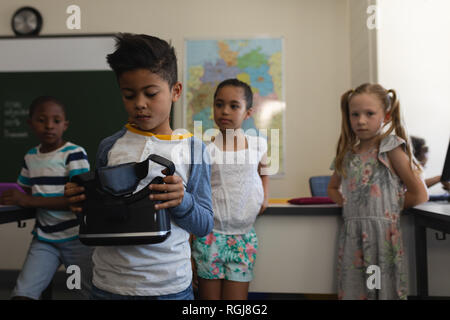 Front view of schoolboy holding and looking at virtual reality headset with his classmates who look him behind  in classroom of elementary school Stock Photo