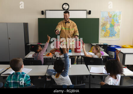 Front view of a schoolkids raising hands while male Caucasian firefighter teaching about fire safety in classroom of elementary school Stock Photo