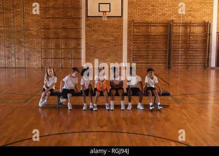 Front view of schoolkids with basketball sitting on bench at basketball court Stock Photo