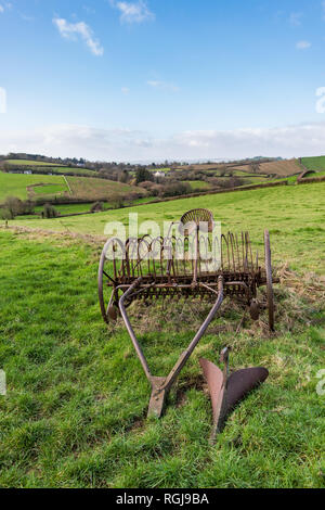 Old rusty horse drawn hay rake in a grass field with Devonshire countryside and hills in the background under a blue and cloudy sky on a bright day Stock Photo