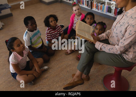 High angle view of female teacher sitting on chair and reading a story schoolkids sitting on floor in school library Stock Photo