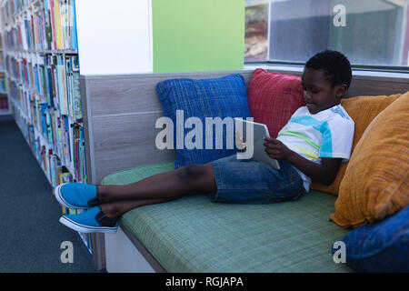 Schoolboy playing on digital tablet while sitting on couch in school library Stock Photo