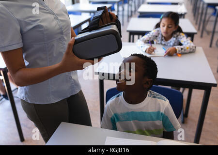 Mid section of female teacher wearing virtual reality headset to schoolboy at desk in classroom of elementary school Stock Photo