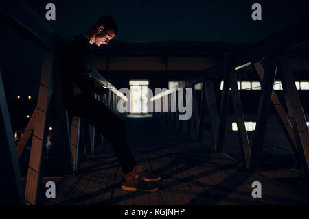 Sportive young man with smartphone and earphones standing on a bridge at night Stock Photo