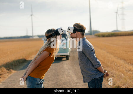 Young couple wearing VR glasses kissing at camper van in rural landscape Stock Photo