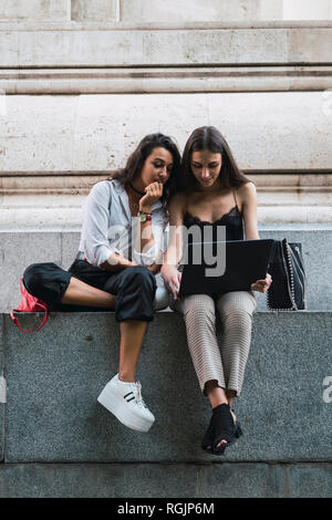 Two friends sitting side by side on a wall using laptop Stock Photo