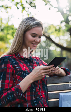 Smiling young woman sitting on bench outdoors using mini tablet Stock Photo