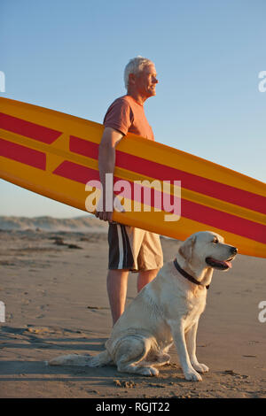 Thoughtful senior man standing on a beach with a surfboard and his dog. Stock Photo
