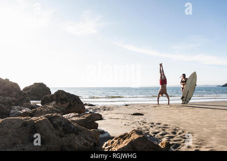 France, Brittany, young man doing a handstand and woman holding surfboard on the beach