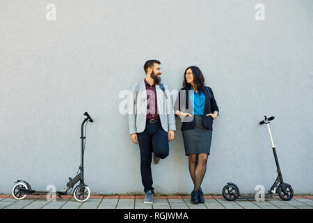 Smiling businessman and businesswoman with scooters leaning against a wall Stock Photo