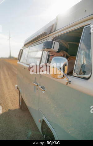 Happy woman wearing VR glasses leaning out of window of a camper van Stock Photo
