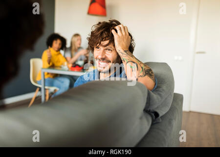 Happy couple sitting on couch talking with friends in background Stock Photo
