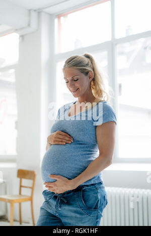 Smiling pregnant woman standing at the window looking at her belly Stock Photo