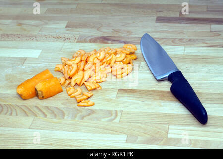 Sliced carrots and a large steel kitchen knife with black handle lying on brown cutting board. Closeup view Stock Photo
