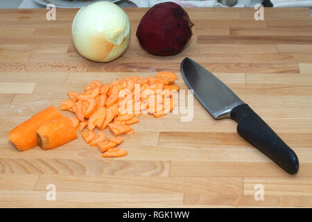 Sliced carrots, whole beets, onions and a large kitchen knife lying on brown cutting board in the kitchen Stock Photo