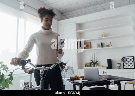 Mid adult woman leaving her home office, pushing bicycle, using smartphone Stock Photo
