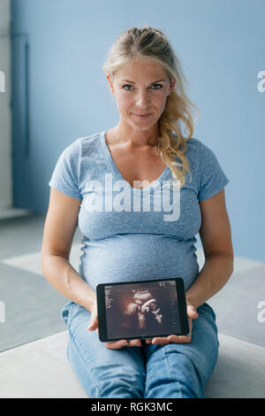 Portrait of smiling pregnant woman sitting on the floor showing ultrasound picture on tablet Stock Photo