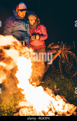 Couple with with bikes standing at camp fire looking at flame by night Stock Photo