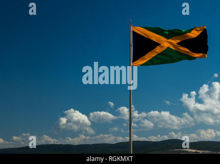 A large Jamaican flag fling in a stiff coastal breeze with blue sky in the background. Flag of Jamaica high above the port city of Falmouth. Stock Photo