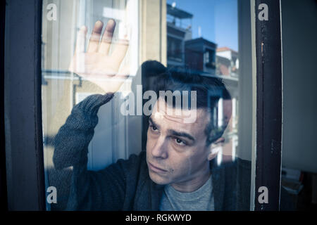 Close up portrait of young sad unhappy caucasian man looking worried, desperate and suicidal through the window at home. feeling worthless and in pain Stock Photo