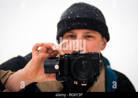 Focus on the camera of a caucasian young man model, photography amateur, filming outside, in cold snowy/rainy weather. Facing the camera, concentrated Stock Photo