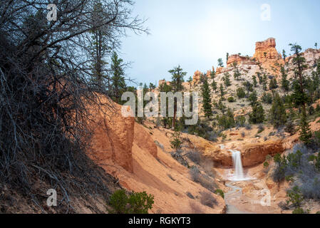 Landscape scene of waterfall on Moss Cave Trail featuring creek and mountains in background framed by gnarled branches in foreground Stock Photo