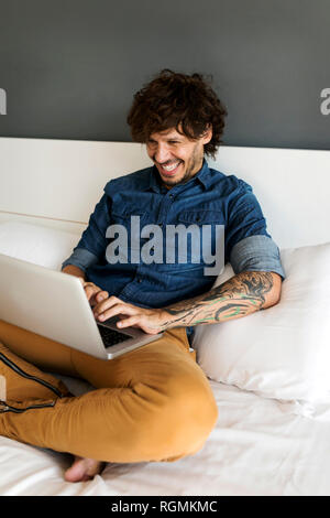 Happy tattooed man sitting on bed using laptop Stock Photo