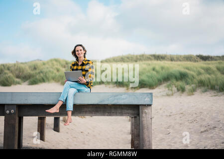 Mature woman sitting on boardwalk at the beach, using laptop Stock Photo