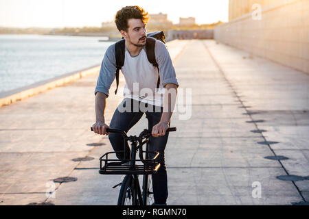 Young man with backpack riding bike on waterfront promenade at the riverside Stock Photo