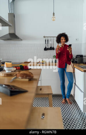 Woman drinking white wine in her kitchen, using smartphone Stock Photo