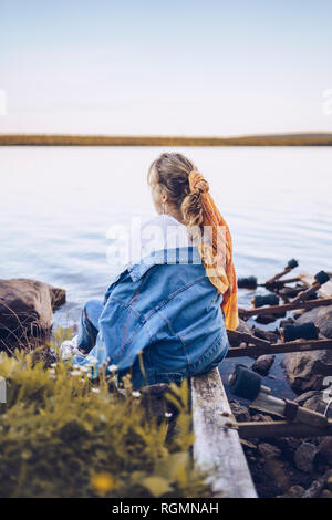 Sweden, Lapland, back view of young woman sitting at water's edge looking at distance Stock Photo