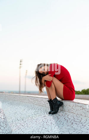 Spain, Barcelona, Montjuic, young woman wearing red dress sitting on a wall Stock Photo
