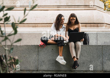 Two friends sitting side by side on a wall using laptop Stock Photo