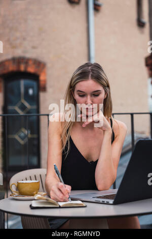 Portrait of young woman sitting on balcony with laptop taking notes Stock Photo