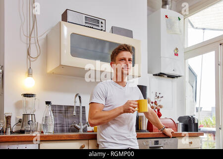 Smiling young man at home having a coffee break in kitchen