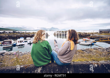 Norway, Senja, two young woman sitting at a small harbor with cell phone