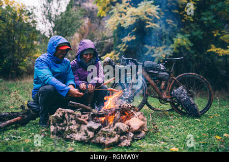 Laughing couple with with bmx bikes sitting at camp fire grilling sausages Stock Photo