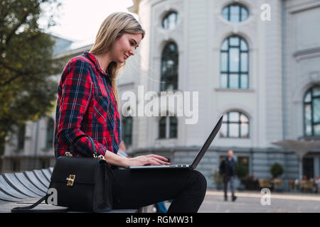 Fashionable young woman sitting on bench outdoors working on laptop Stock Photo
