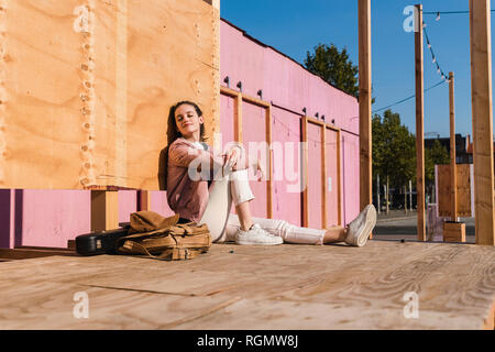 Relaxed young woman sitting on platform next to guitar case and bag Stock Photo