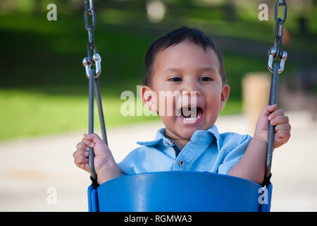 2 year old boy yelling out loud while riding a blue swing at a local park. Stock Photo