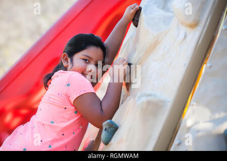 Young latino girl climbing a playground wall and smiling. Stock Photo