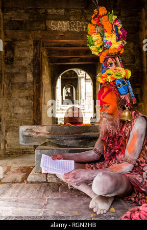 A colorfully decorated Sadhu, Holy Man, reading a book at the ghats of Bagmati river at Pashupatinath Temple Stock Photo