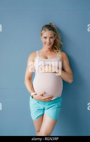 Portrait of smiling pregnant woman standing at blue wall Stock Photo
