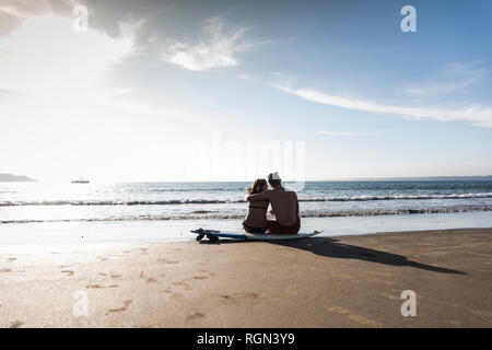 France, Brittany, rear view of young couple sitting on surfboard on the beach