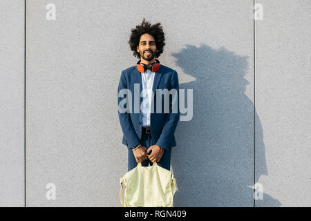 Portrait of smiling young businessman with bag and headphones at a wall Stock Photo