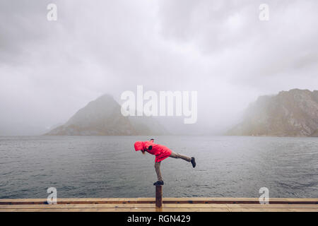 Norway, Lofoten, Hamnoy, man wearing red rain jacket balancing on wooden stake Stock Photo