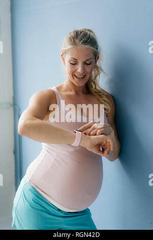 Portrait of smiling pregnant woman standing at blue wall looking at smartwatch Stock Photo