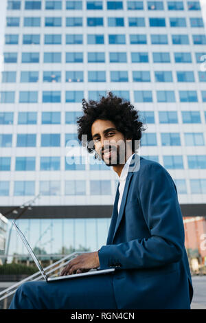 Spain, Barcelona, smiling young businessman sitting outdoors in the city working on laptop Stock Photo